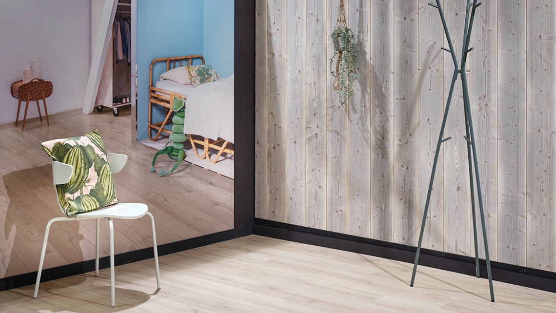 Espace de vie avec une chaise blanche et un porte-manteau. Le mur est en bois clair, et on peut voir une chambre avec un lit à travers une ouverture dans le mur. Des plantes suspendues ajoutent une touche de verdure à la pièce.